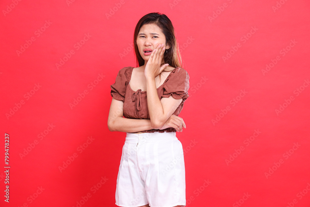 indonesia woman's gesture towards the camera holding her cheek with a sore tooth, wearing a brown blouse and shorts with a red background. for health, care and advertising concept indonesia woman's gesture towards the camera holding her cheek with a sore tooth, wearing a brown blouse and shorts with a red background. for health, care and advertising concept