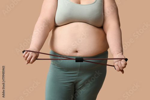 Young overweight woman with fitness expander on brown background, closeup. Weight loss concept