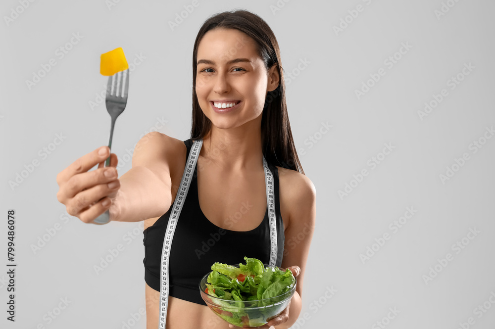 Beautiful young happy woman with measuring tape and bowl of fresh salad on grey background. Weight loss concept Beautiful young happy woman with measuring tape and bowl of fresh salad on grey background. Weight loss concept