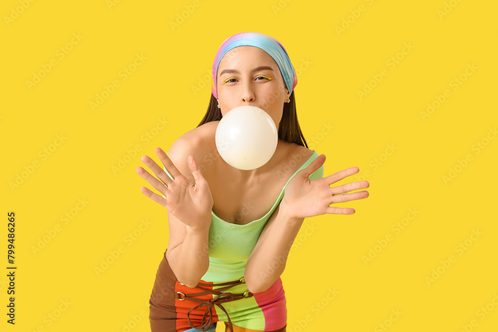 Beautiful young fashionable woman blowing bubblegum on yellow background