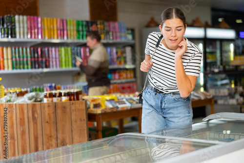 Young attractive woman in casual jeans and t-shirt shopping for groceries in food store, looking at products in glass refrigerator with interest © JackF