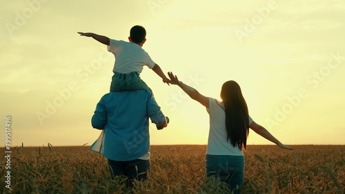 Boy on dad shoulders dad, mom play together in wheat field. Son on dad shoulders, mom holding son hand, family walk in nature. Family agricultural business. Father mother child traveling together
