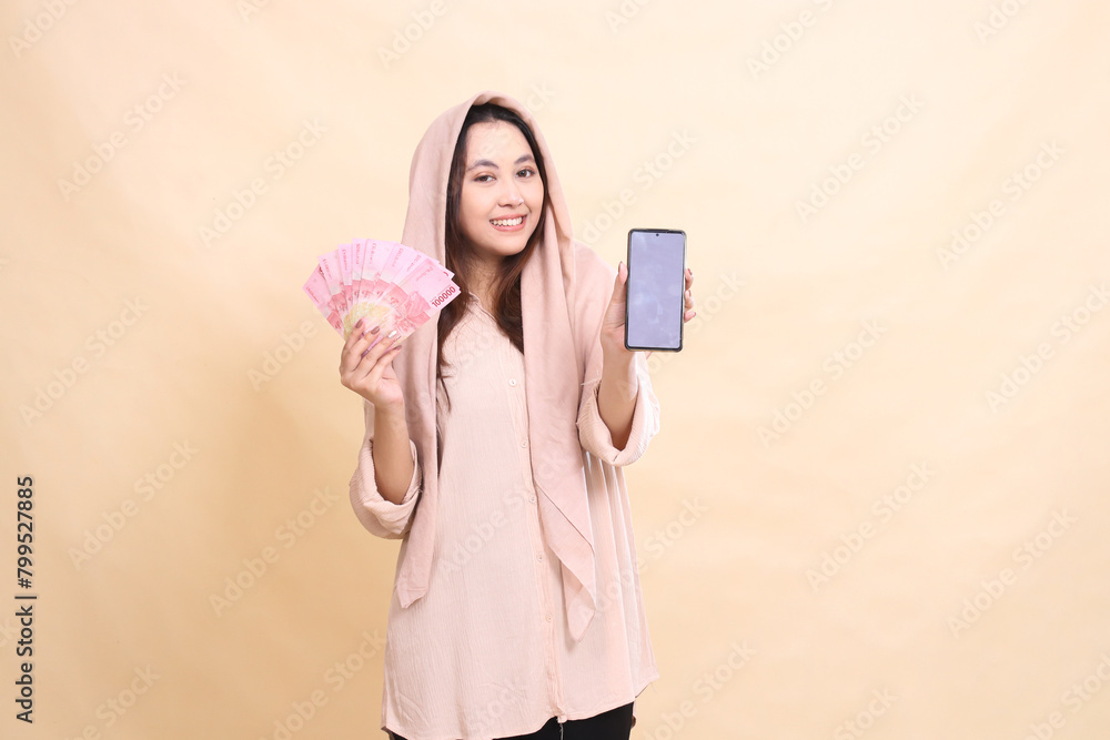 Asian woman smiles at the camera, holding rupiah money and holding the cellphone gadget screen in front of her. beautiful young for finance, technology, fashion Asian woman smiles at the camera, holding rupiah money and holding the cellphone gadget screen in front of her. beautiful young for finance, technology, fashion