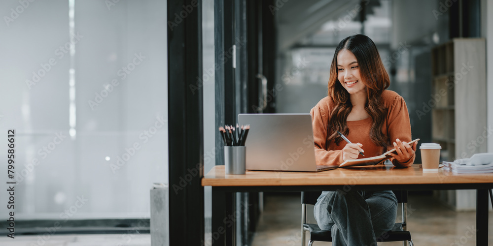 Smiling young woman taking notes while working on her laptop in a well-lit, modern office space. Smiling young woman taking notes while working on her laptop in a well-lit, modern office space.