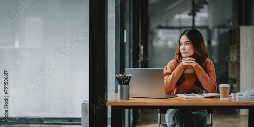 Young businesswoman with hands clasped, sitting at her desk in a modern office, deep in thought. photo