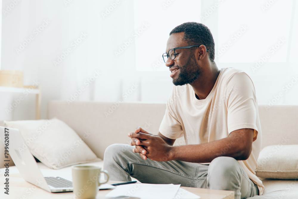 Smiling African American Freelancer Working on Laptop in Modern Home Office A young African American man with a casual style is sitting on a comfortable sofa in a modern living room He is happily