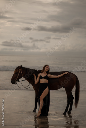 Brown horse and a woman in black dress stand on a beach, with the sky and sea creating a picturesque backdrop for the scene, sunset time