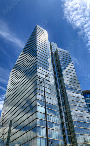 Dominating the frame, a modern skyscraper ascends towards the clear blue sky, lined with fluffy white clouds. Its glass facade reflects the surrounding environment, creating a play of light and shadow