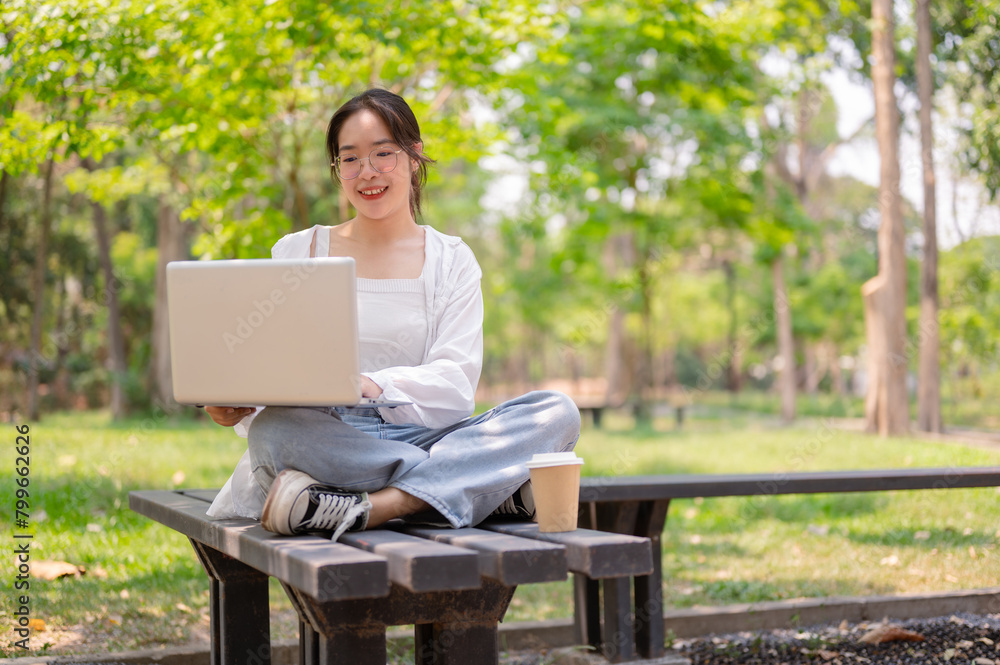 A young beautiful, positive Asian woman sits on a bench in a green park using her laptop computer. A young beautiful, positive Asian woman sits on a bench in a green park using her laptop computer.