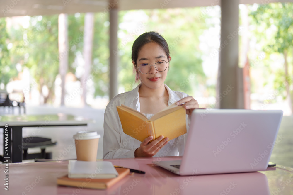A young Asian female college student reading a book while sitting at a table in a campus cafeteria. A young Asian female college student reading a book while sitting at a table in a campus cafeteria.