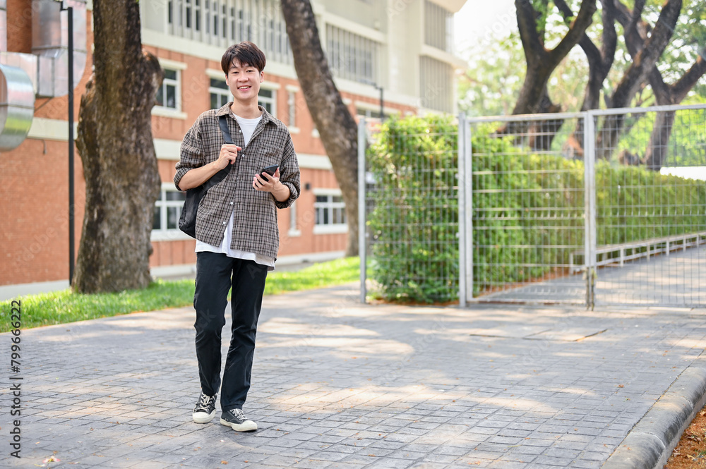 A happy Asian male college student is using his smartphone while walking in the campus park.