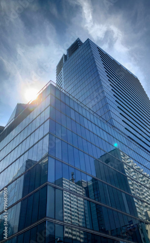 The image captures the imposing architecture of a modern skyscraper. Its reflective glass facade glints with the sun s rays  creating a lens flare effect. The blue sky  peppered with clouds  contrasts