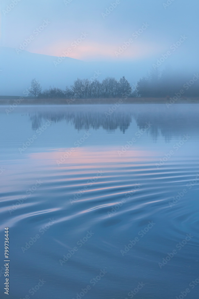 The gentle, rhythmic pattern of waves on a tranquil lake, with the water's surface reflecting the soft hues of the sky at dawn. 