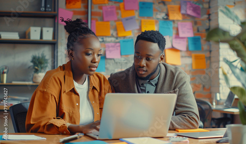 A black female business manager assists a young male adult executive working on a laptop in a modern office. A diverse team of creative workers uses technology for project planning and marketing strat photo