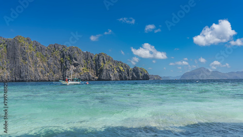 The traditional Filipino double-outrigger dugout bangka boat is anchored in the turquoise ocean. Picturesque steep karst cliffs against a blue sky and clouds. An island in the distance. Philippines. 