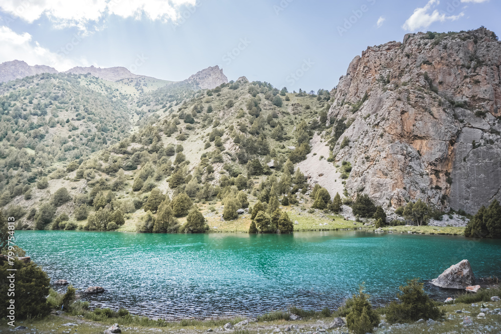 Mountain panorama, landscape with rocky peaks and blue turquoise lake Chukurak in the Fan Mountains in Tajikistan, hills covered with forest on a sunny summer day