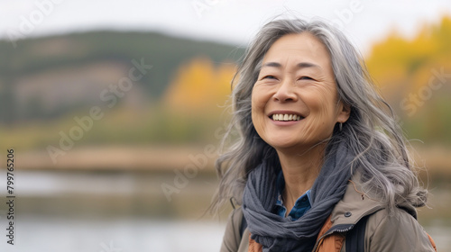 A pretty Asian woman over 50 on a walk in the park or in the forest against the background of a lake in autumn. An optimistic and positive woman aged in nature.