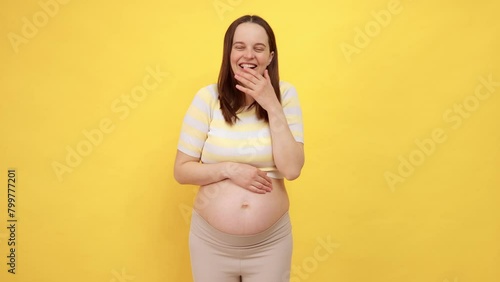 Satisfied delighted positive Caucasian pregnant woman with bare belly wearing casual top isolated over yellow background laughing loud stroking her belly having fun
