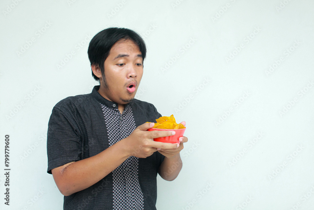 A young asian man with a surprised expression holds a bowl of potato snacks. Isolated in gray background. A young asian man with a surprised expression holds a bowl of potato snacks. Isolated in gray background.