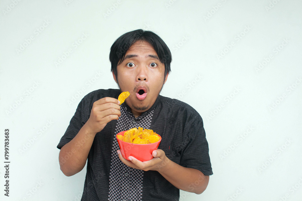 A young asian man with a surprised expression holds a bowl of potato snacks. Watch film concept. Isolated in gray background. A young asian man with a surprised expression holds a bowl of potato snacks. Watch film concept. Isolated in gray background.