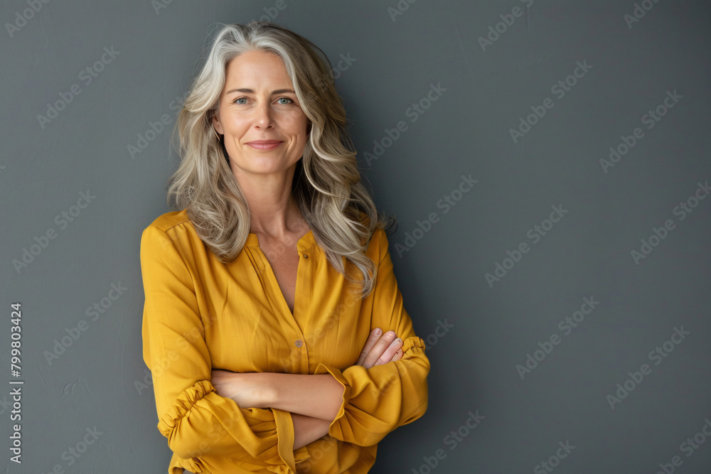 Portrait of a beautiful smiling middle aged woman standing with her arms crossed on a grey background, looking at the camera, wearing a yellow blouse and casual outfit Portrait of a beautiful smiling middle aged woman standing with her arms crossed on a grey background, looking at the camera, wearing a yellow blouse and casual outfit