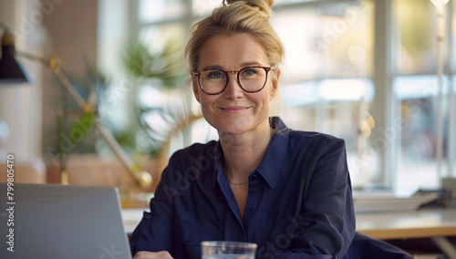 Happy businesswoman working on laptop in office . young woman with blonde hair wearing blue shirt and glasses sitting at desk using computer while smiling to camera 