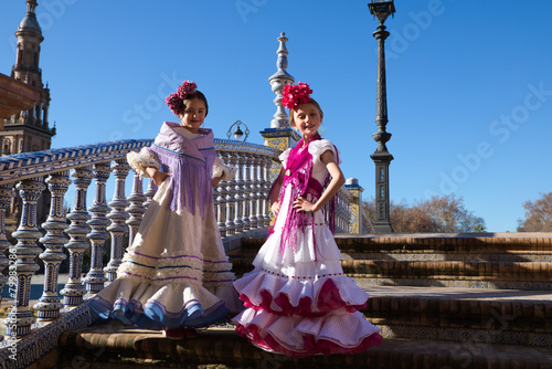 Two pretty little girls dancing flamenco dressed in typical gypsy costume pose on the stairs in their dress in a famous square in seville. The girls are happy. Flamenco, cultural heritage of humanity.