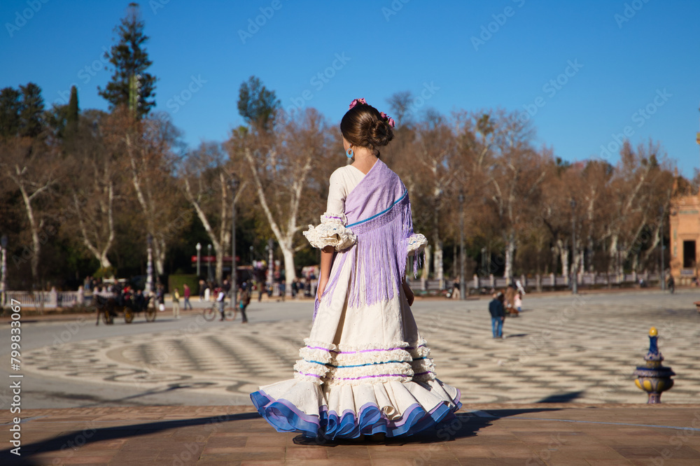 a little girl dancing flamenco dressed in a beige dress with ruffles and purple fringes in a famous square in seville, spain. The girl has flowers on her head and her hair in a bun. a little girl dancing flamenco dressed in a beige dress with ruffles and purple fringes in a famous square in seville, spain. The girl has flowers on her head and her hair in a bun.