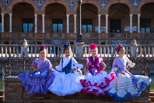 Four little girls dancing flamenco dressed in typical flamenco dress sitting on a bench in a famous square in seville, spain. In the background the arches and columns of the famous square.