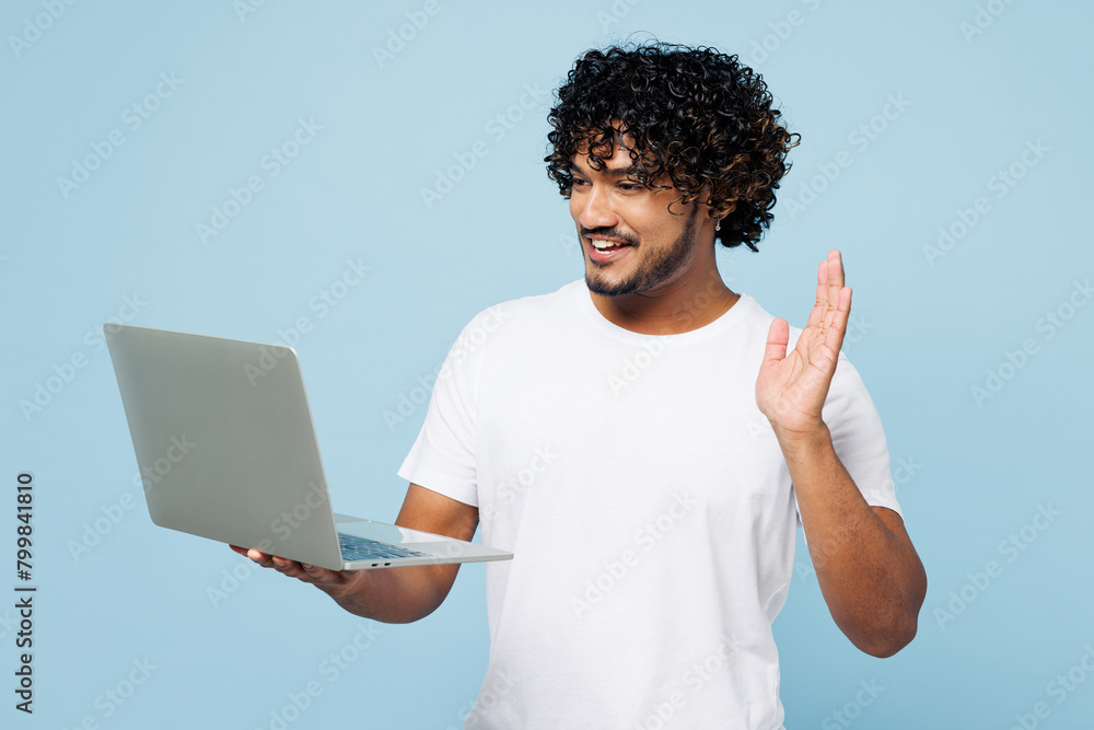 Young IT Indian man he wear white t-shirt casual clothes hold use work on laptop pc computer waving hand talk speak by video call isolated on plain pastel light blue cyan background Lifestyle concept Young IT Indian man he wear white t-shirt casual clothes hold use work on laptop pc computer waving hand talk speak by video call isolated on plain pastel light blue cyan background Lifestyle concept