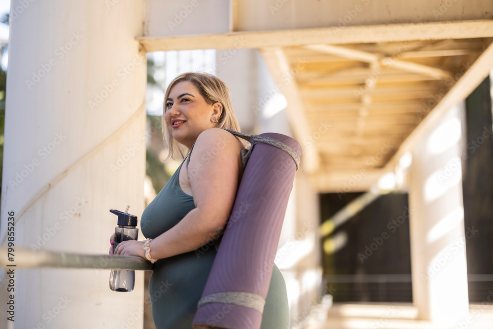 Smiling young curvy woman standing with exercise mat and water bottle Smiling young curvy woman standing with exercise mat and water bottle