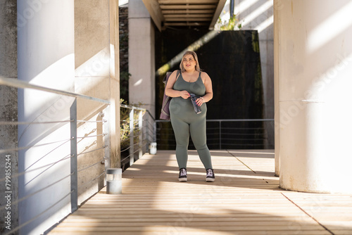 Smiling curvy woman standing with water bottle near railing photo