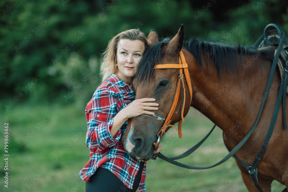 Obraz premium Happy blonde with horse in forest. Woman and a horse walking through the field during the day. Dressed in a plaid shirt and black leggings.