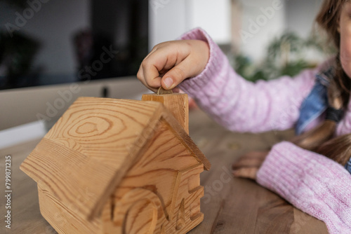 Girl inserting coin into wooden house shape bank at table at home photo