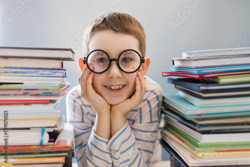 Smiling boy wearing eyeglasses leaning near stacks of books at home photo