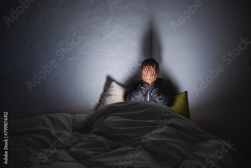 Depressed man covering face with hands sitting on bed in dark room