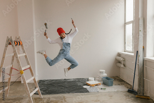 Cheerful woman with paint equipment jumping in front of white wall at new home
