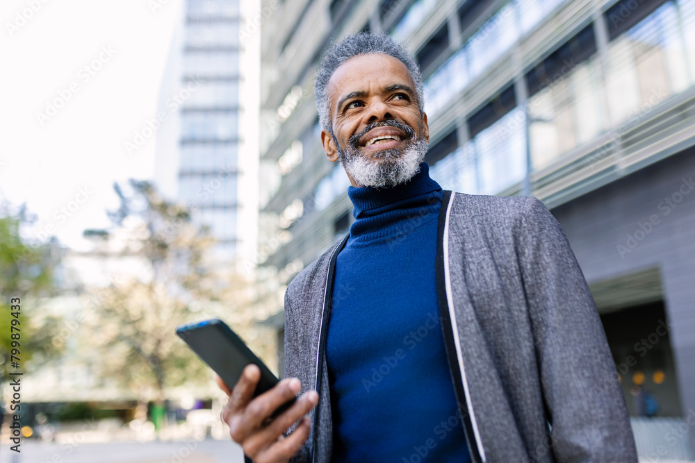 © Westend61 - Happy businessman standing with smart phone in front of building