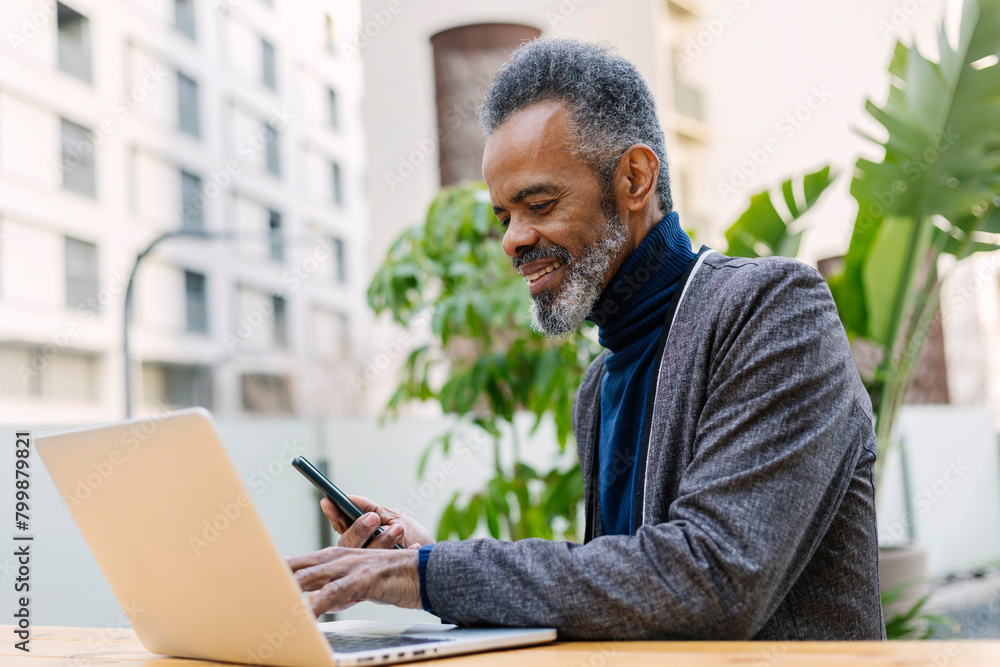 Smiling mature businessman holding smart phone and working on laptop