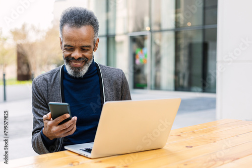 Smiling businessman using smart phone and sitting with laptop at table photo