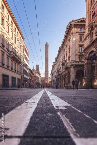 A low-angle perspective of a Bologna street, emphasizing the road lines converging towards the Asinelli and Garisenda Towers. The architecture on either side is illuminated by warm sunlight