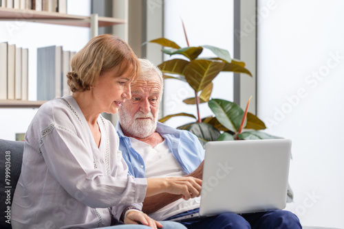 Senior couple checking their bills, man and a woman Using laptop on a cozy sofa at home, retired elderly family reading documents
