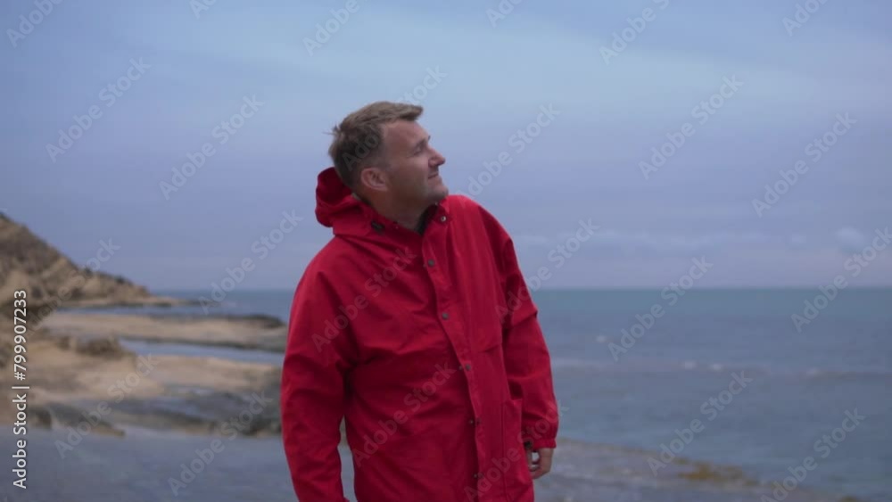 Handsome brunette man in red jacket looking at overcast sky on a cliff by the sea. Handsome brunette man in red jacket looking at overcast sky on a cliff by the sea.