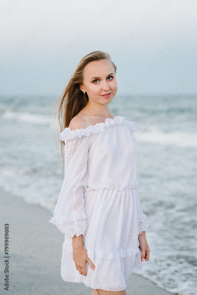 Portrait of a beautiful girl in a white dress. Female stand on the beach ocean and enjoy summer day on vacation. Happy woman go on seashore. Girl walks in water with big waves on sand sea in evening. Portrait of a beautiful girl in a white dress. Female stand on the beach ocean and enjoy summer day on vacation. Happy woman go on seashore. Girl walks in water with big waves on sand sea in evening.
