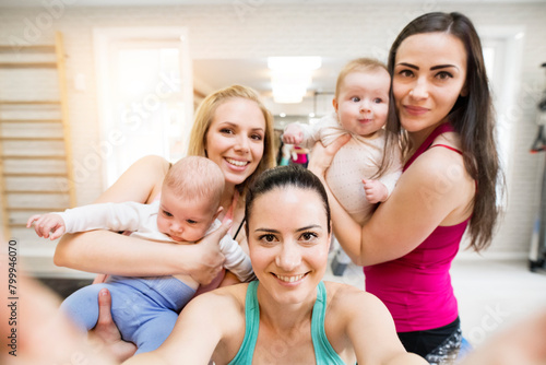 Group exercise class, mothers working out with babies in gym. Friendship between parents taking selfie.