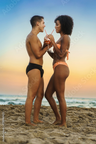 Multiethnic couple sipping a shared cocktail on the beach at sunset - Intimate moment with ocean backdrop - Celebrating love and diversity.
