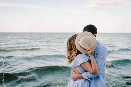 Hat hidden face. Woman and man hiding faces on beach ocean and enjoys sunny summer day. Female and male hide faces behind straw hat and kiss on sand sea. Secret, incognito concept. Couple in love hugs