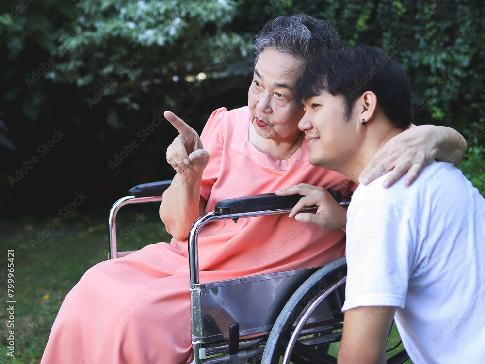 Obraz premium Happy Asian senior woman sitting on wheelchair with her son in the garden. talking and pointing.