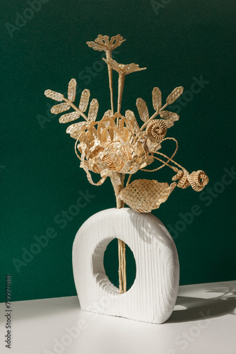 A wooden vase with a bouquet of flowers made from straw on a green background. Straw weaving