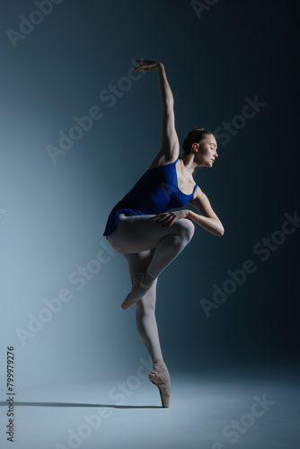 Young ballerina in bodysuit and pointe shoes dancing against of blue background.